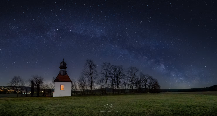 A small chapel stands in a meadow surrounded by a few trees, its windows illuminated by a warm yellow light. The Milky Way stretches in an arc above the chapel.