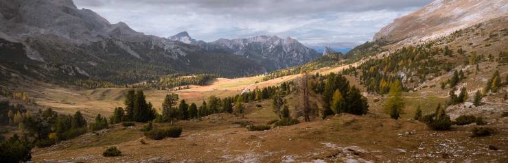 A long valley lined with rugged mountains stretches to the horizon. The valley is marked by larch trees and pastures, which are turning orange-brown as autumn approaches. Parts of the valley are partially illuminated by the sun.