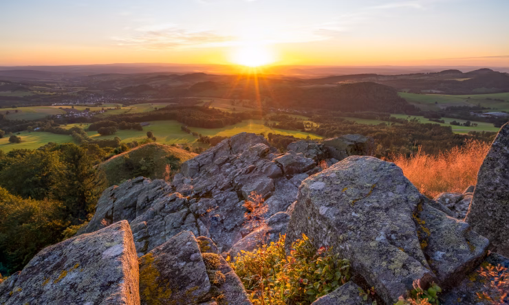 The sun sets over the green hills of the Rhön, bathing everything in a warm light. An orange-lit rock dominates the foreground.