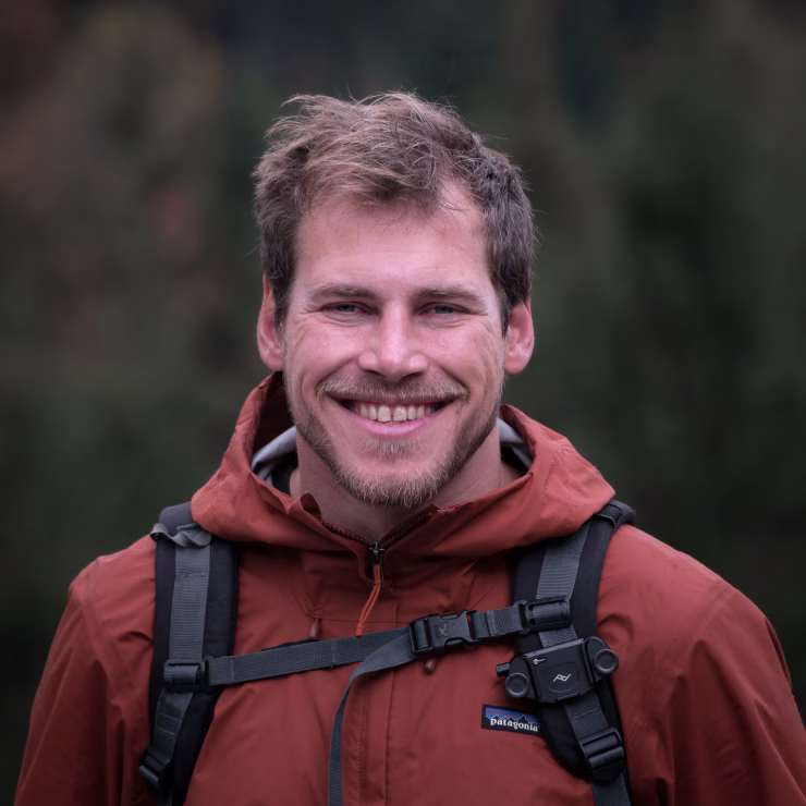 A portrait of Thorsten Zobel, landscape and nature photographer from Frankfurt. The image is taken in front of a forest, wearing a backpack and a rain jacket.